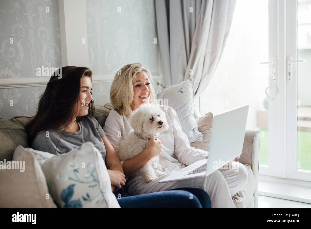 Mother and her daughter video calling someone on their laptop. They are smiling and waving at the screen. Their dog is sitting with them. Stock Photo