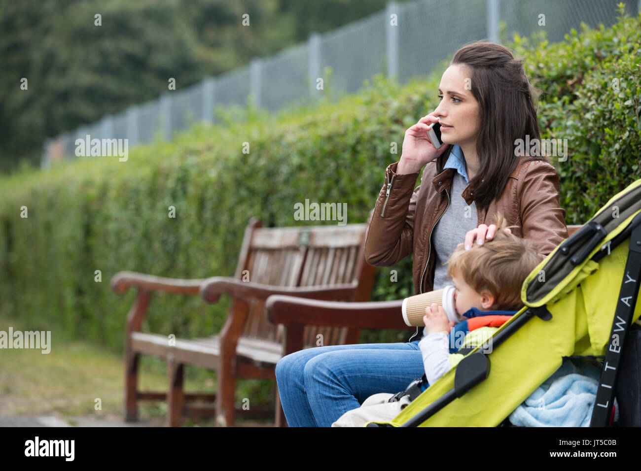 Busy Mother using mobile phone with toddler sitting in pram. Stock Photo