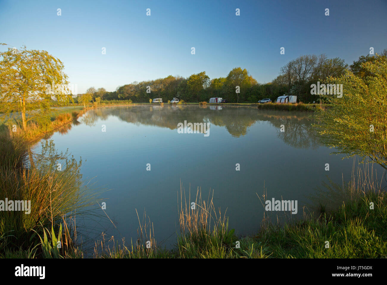 Caravans and campervan beside trees in rural landscape reflected in calm blue water of coarse fishing pond under blue sky in England Stock Photo