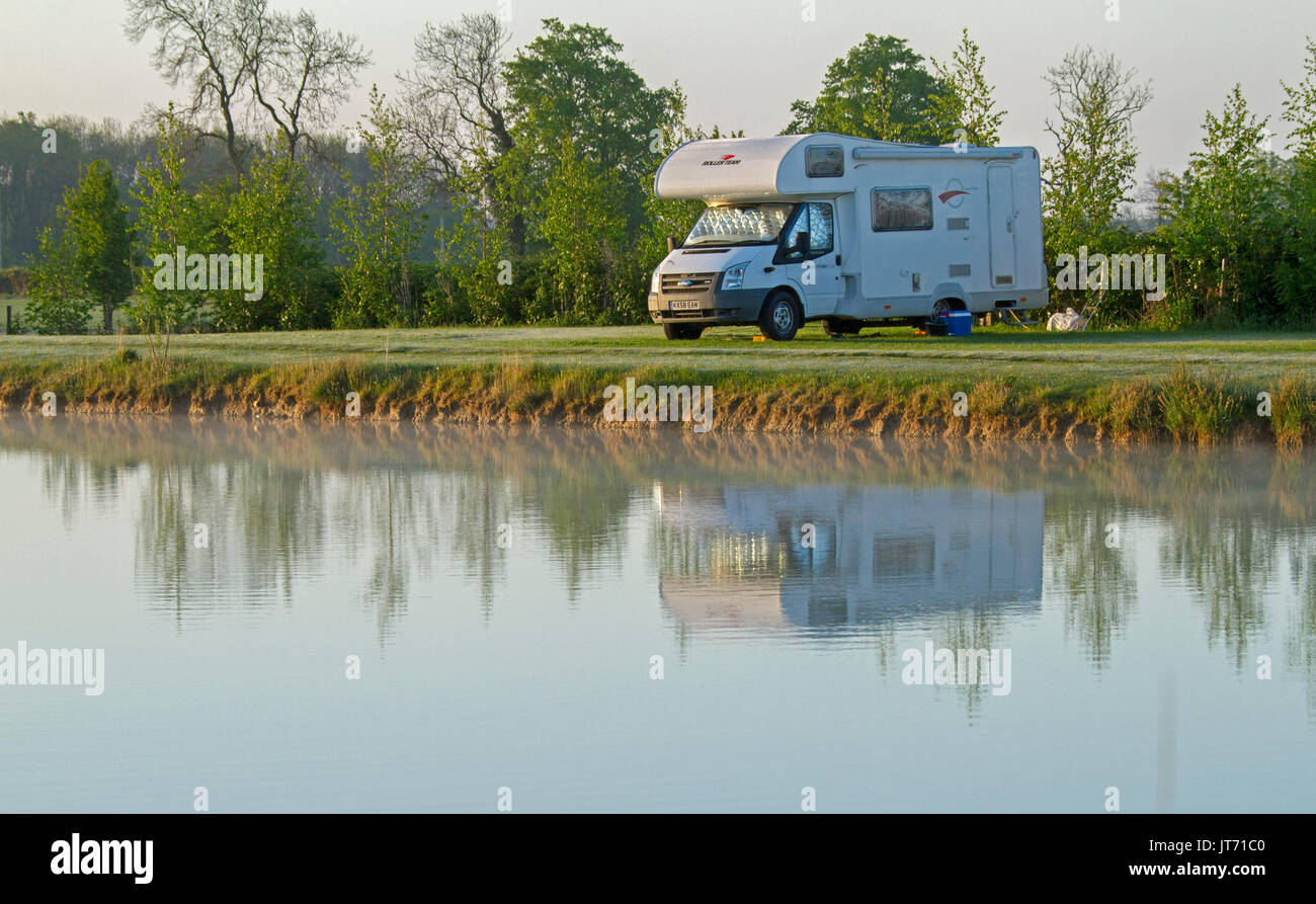 Motorhome reflected in calm water of adjacent coarse fishing pond at dawn at farm camping area in England Stock Photo