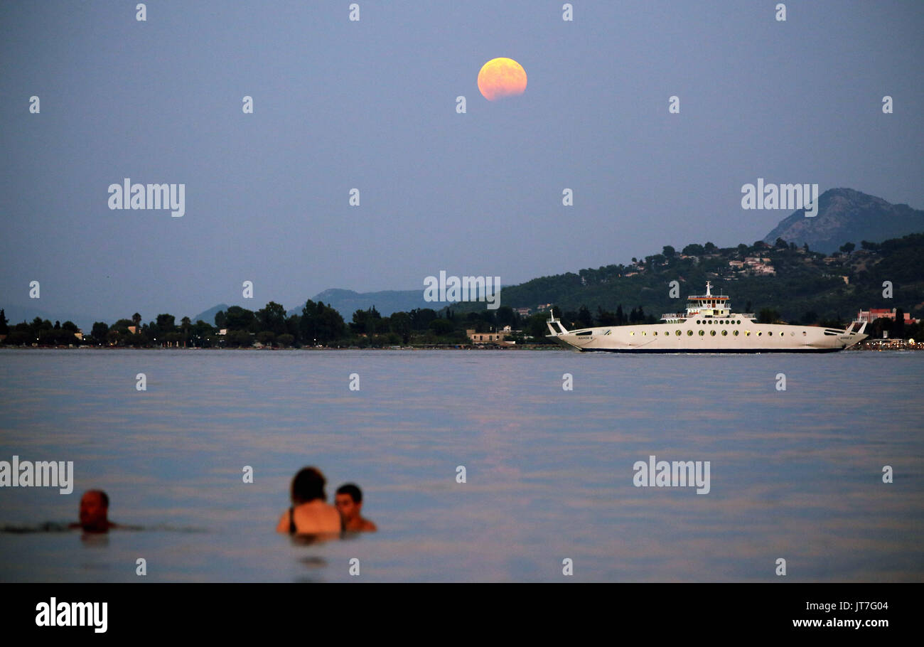 A full moon with a partial eclipse rises, while a ferry boat departs for Eretria and  people cool off in the sea in Oropos in the Evia Bay, 30 miles e Stock Photo