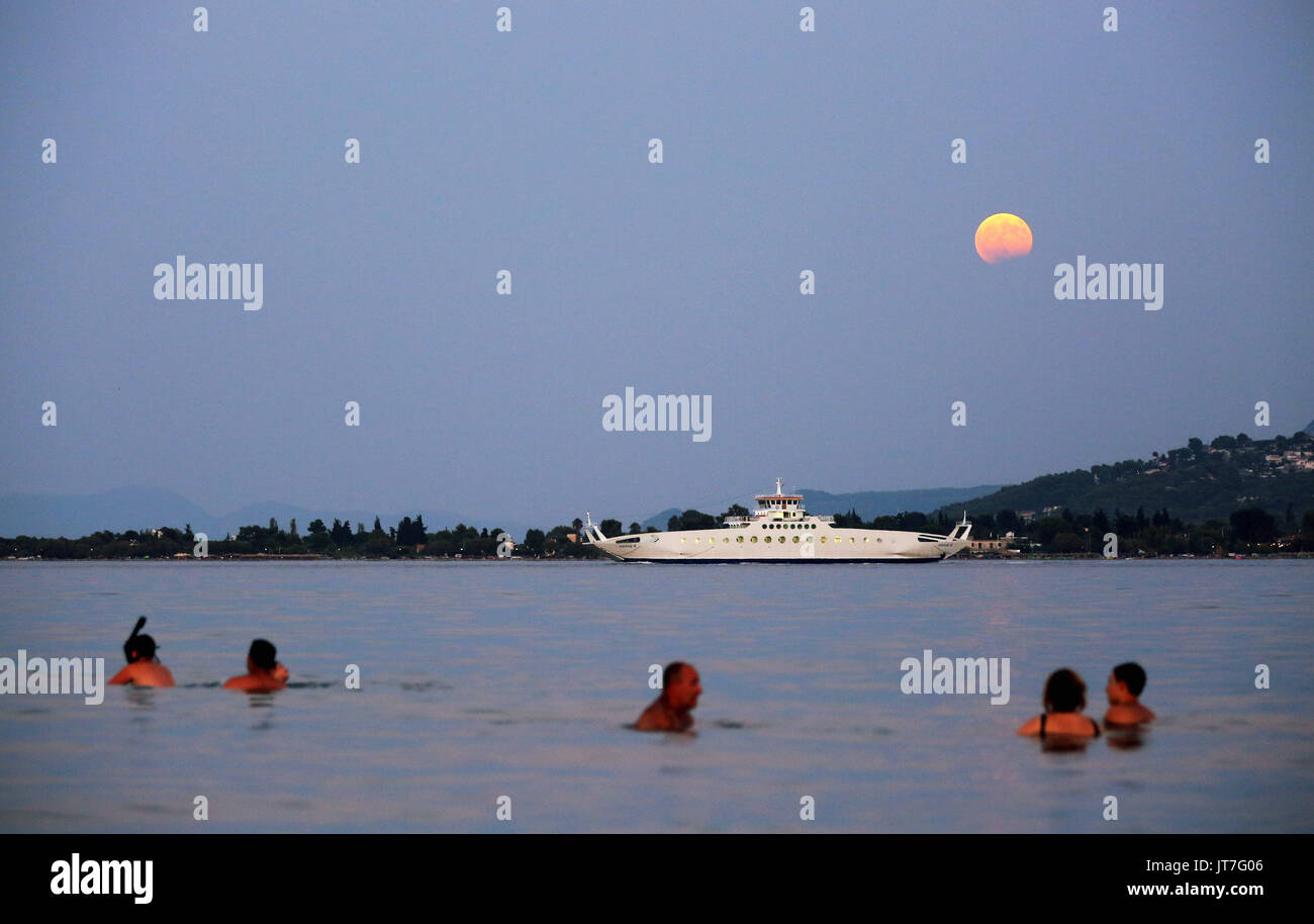 A full moon with a partial eclipse rises, while a ferry boat departs for Eretria and  people cool off in the sea in Oropos in the Evia Bay, 30 miles e Stock Photo