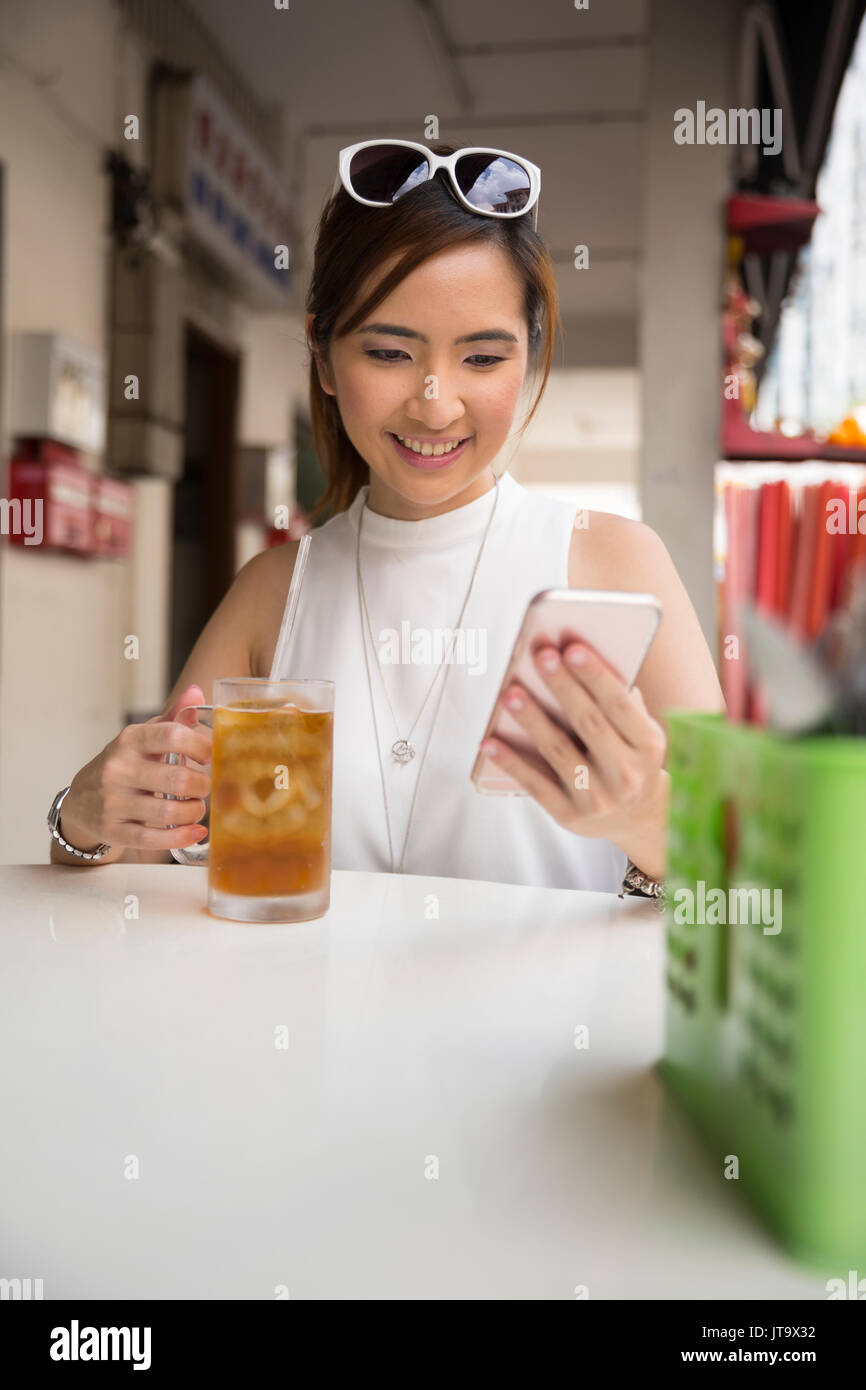 Chinese woman drinking an ice lemon tea, while sitting with her phone in an Asian food court or Hawker centre cafe. Stock Photo