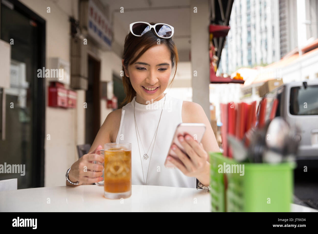 Chinese woman drinking an ice lemon tea, while sitting with her phone in an Asian food court or Hawker centre cafe. Stock Photo