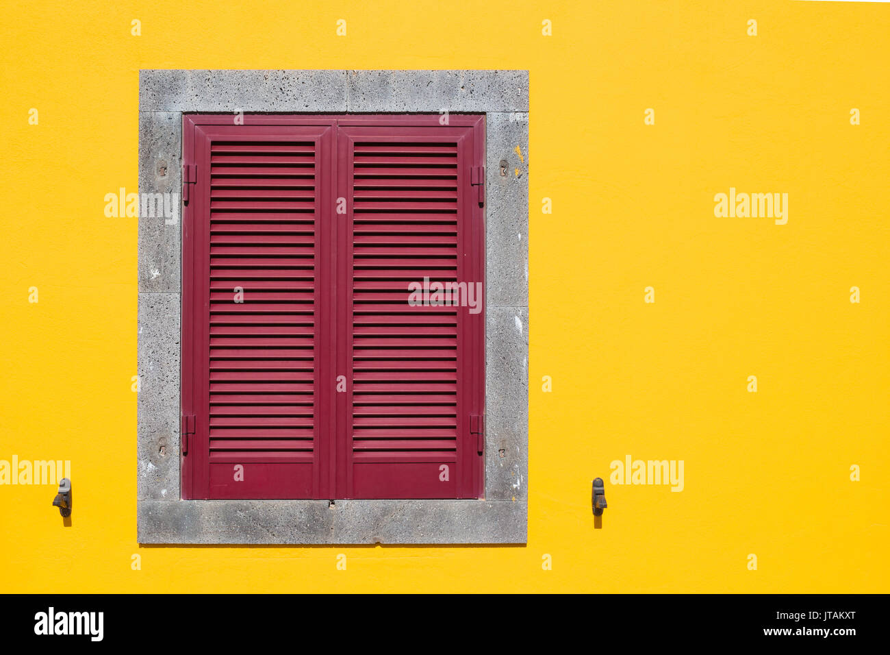 Bright yellow facade with a stone framed grey window, covered with red shutter. Stock Photo