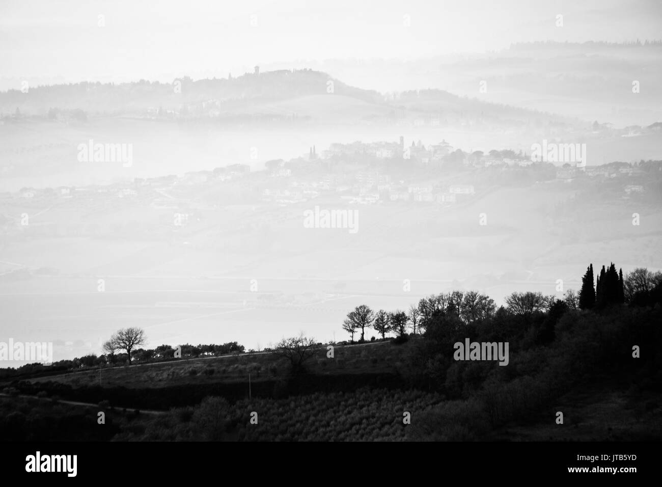 A view of some trees in the foreground with layers of hills and towns through the mist in the background Stock Photo