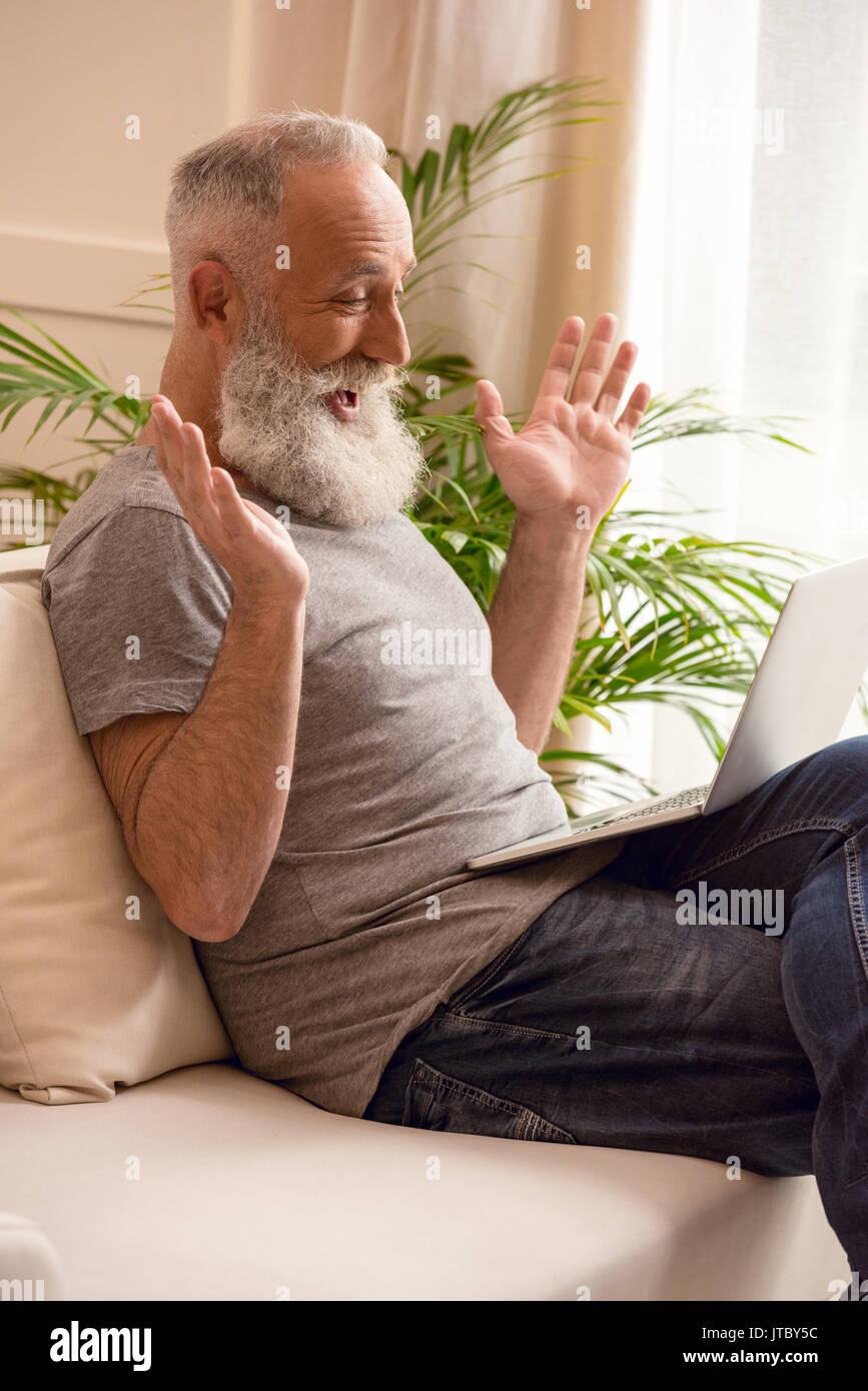 surprised senior bearded man using laptop while sitting on sofa at home Stock Photo