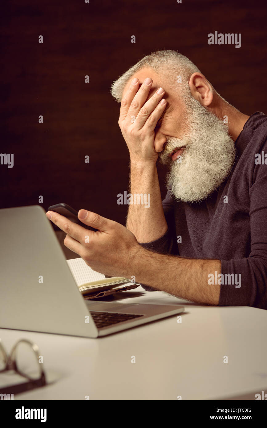 grey haired man sitting at table and holding smartphone with facepalm at home Stock Photo
