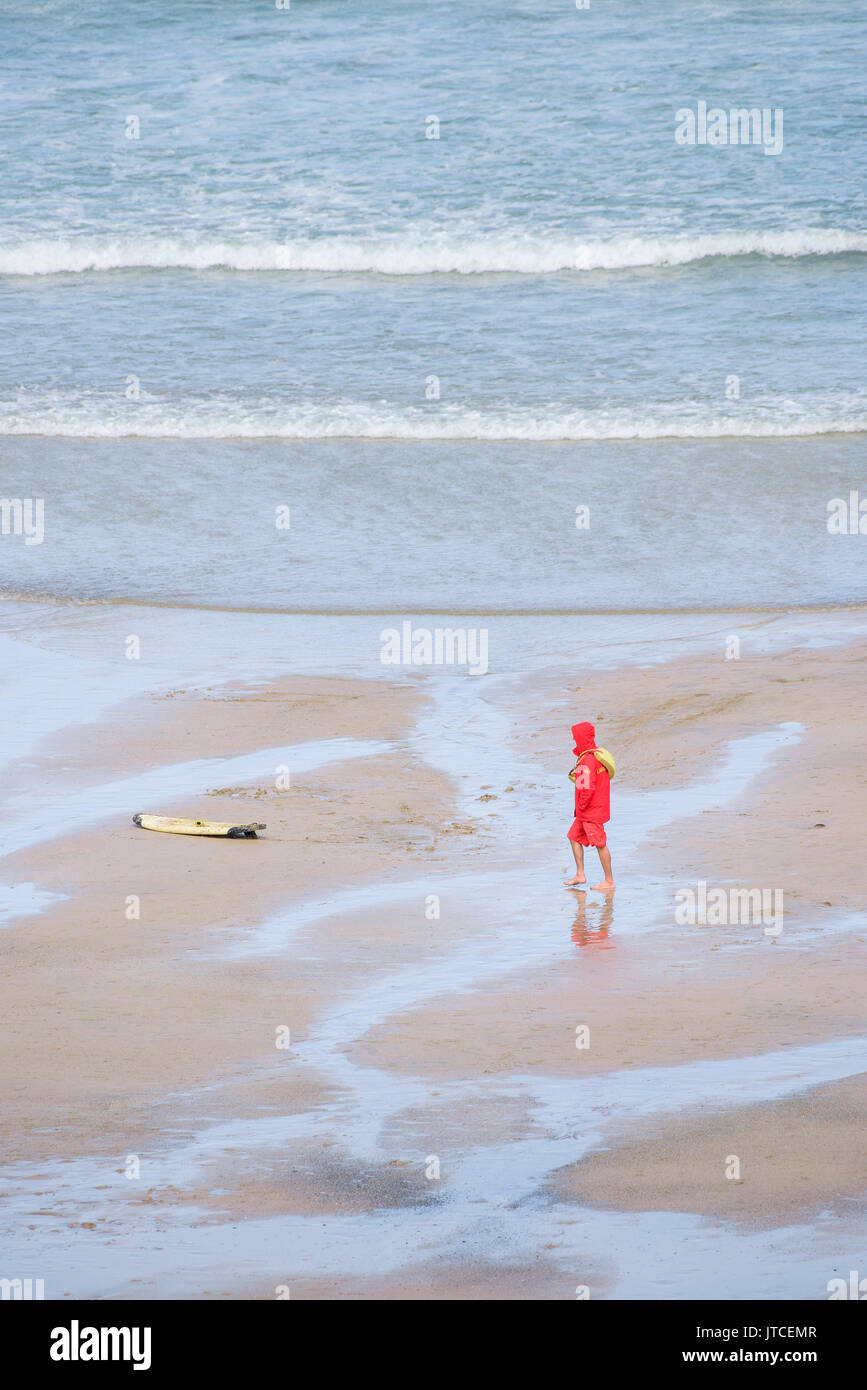 A RNLI Lifeguard on duty walking along the shoreline on a beach in Newquay, Cornwall. Stock Photo
