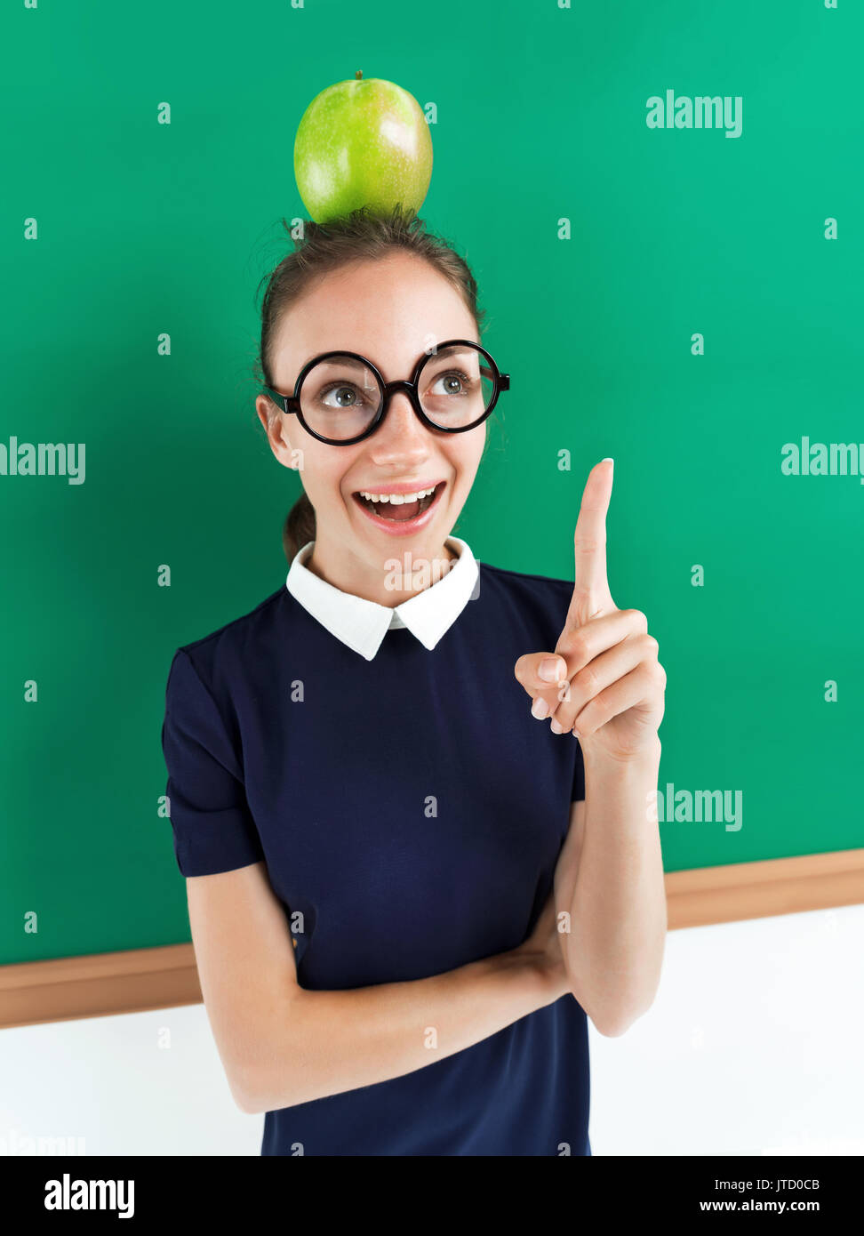 Cheerful enthusiastic pupil with apple on her head pointing finger up. Photo of teen wearing glasses near blackboard, creative concept with Back to sc Stock Photo