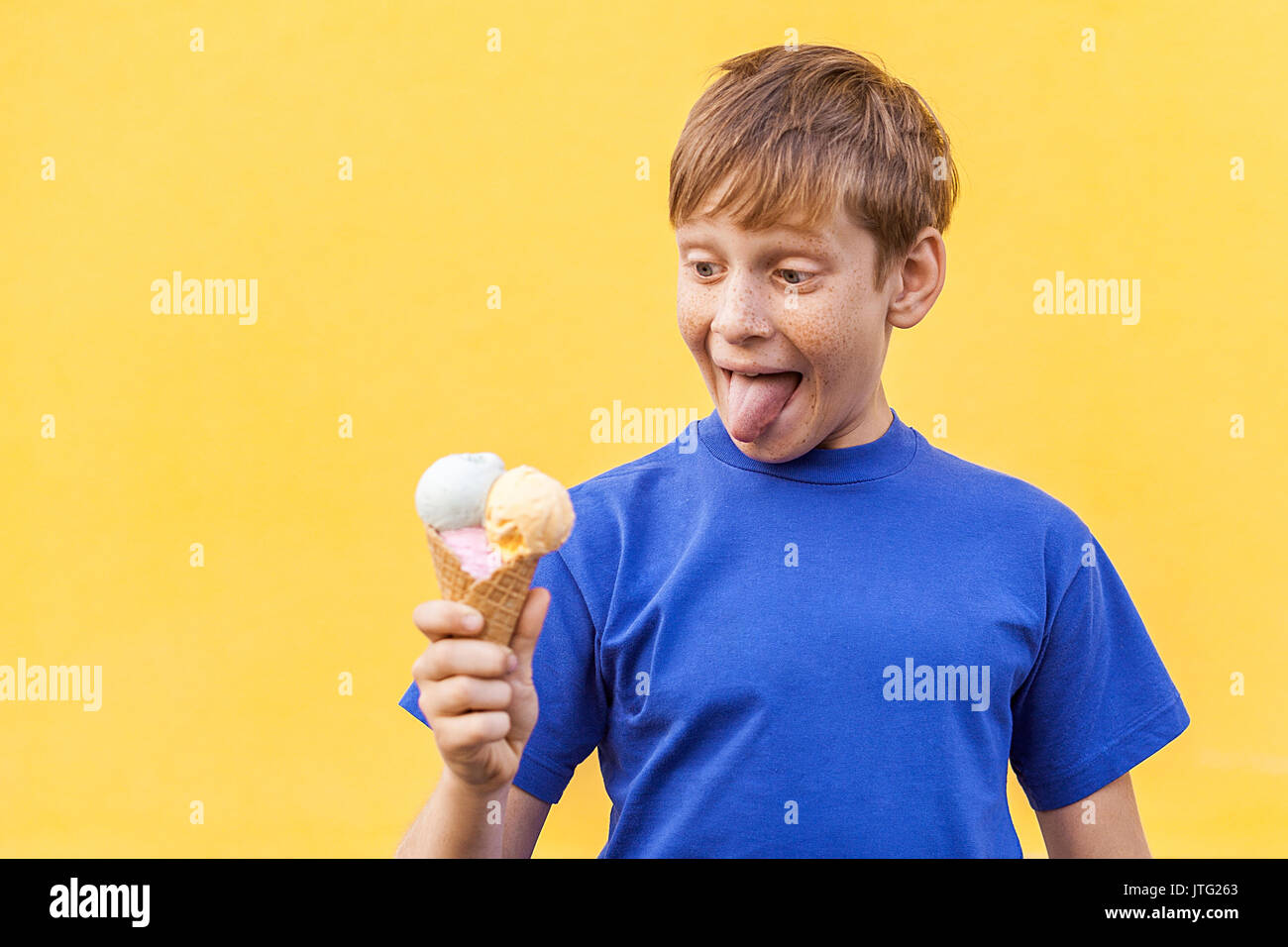 The blondå beautiful boy with freckles and blue t-shirt looking at  ice cream  with surprised face and tongue out. Studio shot, isolated on a yellow b Stock Photo