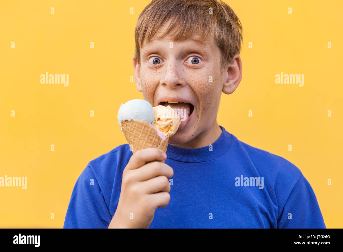Blonde beautiful boy with freckles and blue T-shirt eating and  lickes ice cream  and looking at camera with surprised face. Studio shot, isolated on  Stock Photo
