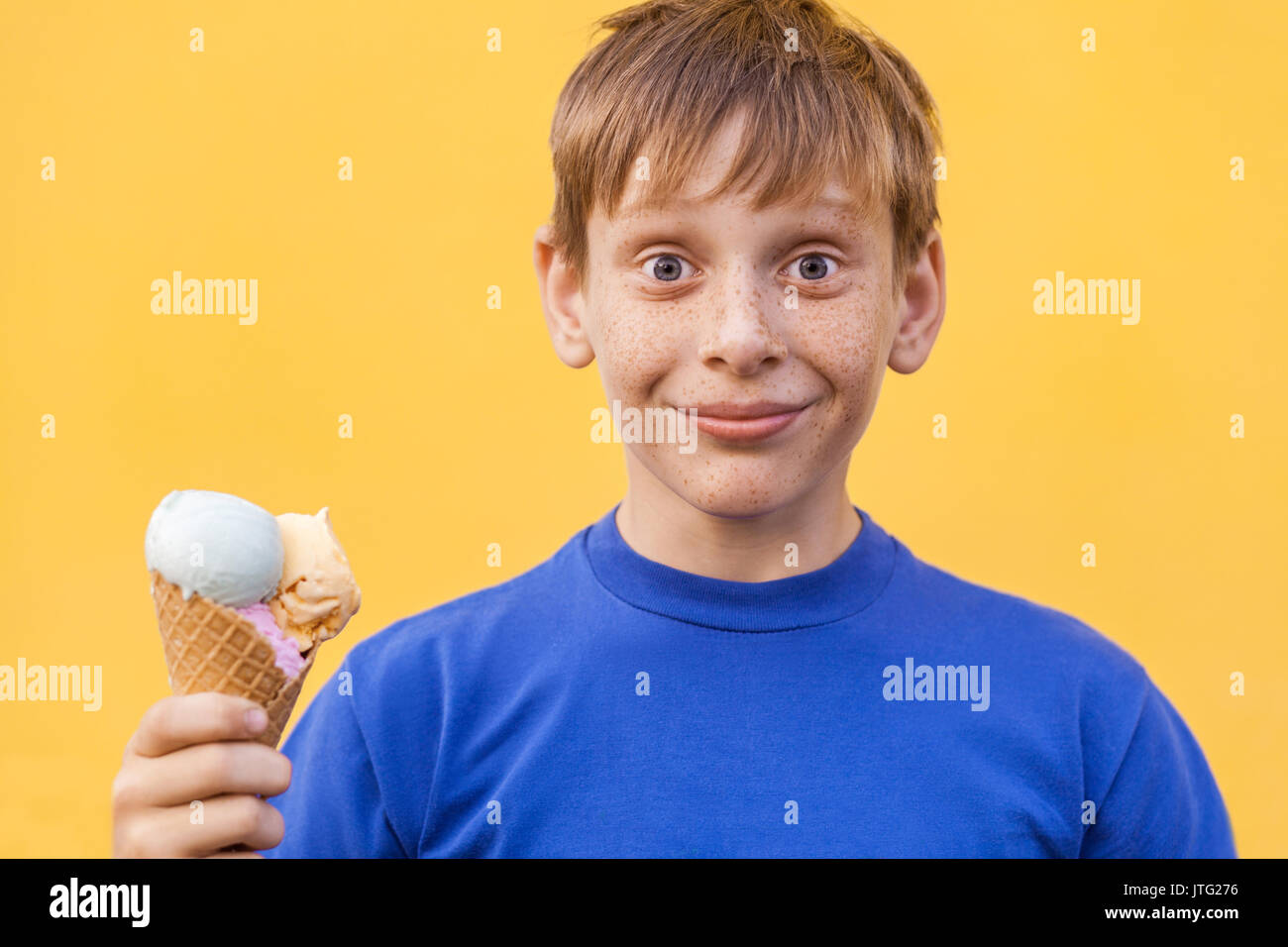 Blonde beautiful boy with freckles and blue t-shirt holds ice cream looking at camera with smile and wonder face . Studio shot, isolated on a yellow b Stock Photo