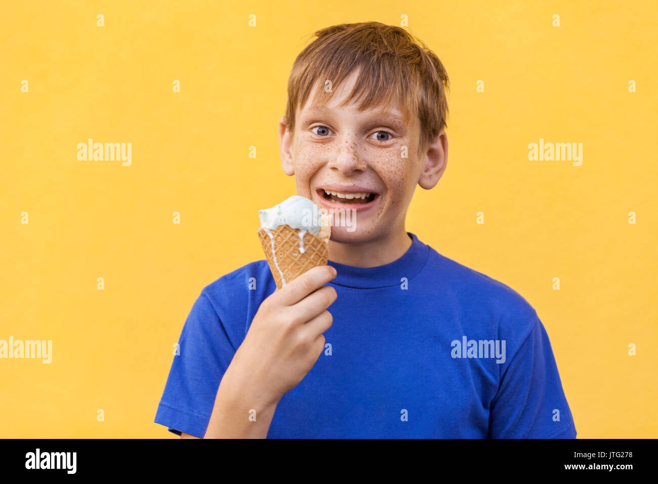 The blonde beautiful boy with freckles and blue T-shirt holds ice cream looking at camera with toothy smile. Studio shot, isolated on a yellow backgro Stock Photo