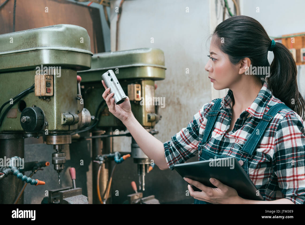 beautiful milling machining employee girl holding mobile digital tablet standing in front of drilling machine and holding components doing inspection. Stock Photo
