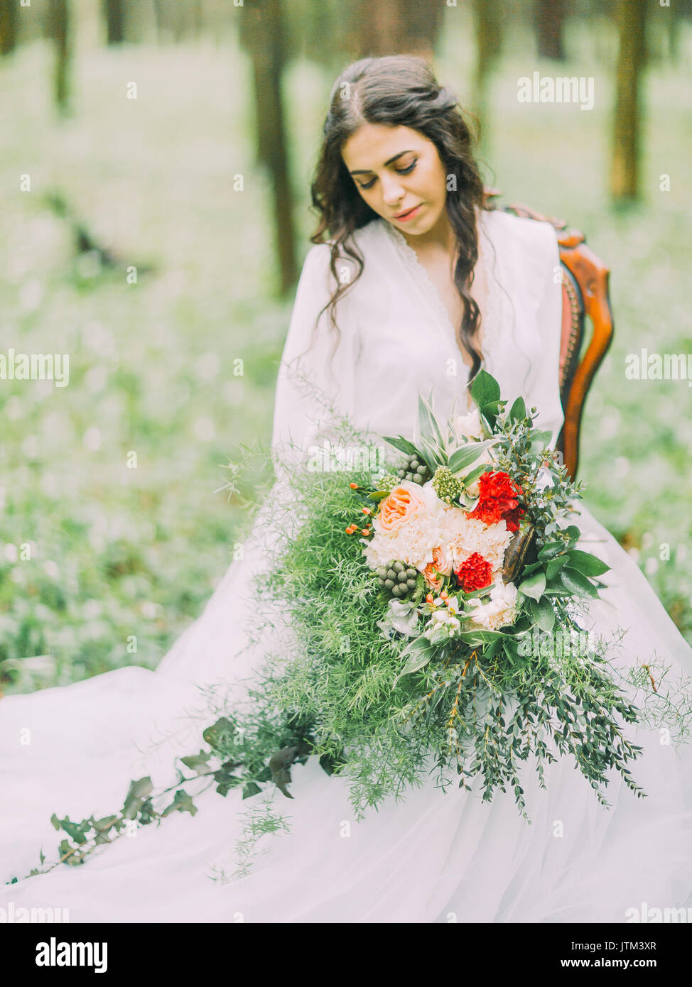 Beautiful bride with brown hair looking at her wedding clourful bouquet and sitting on the chair in the green wood. Stock Photo