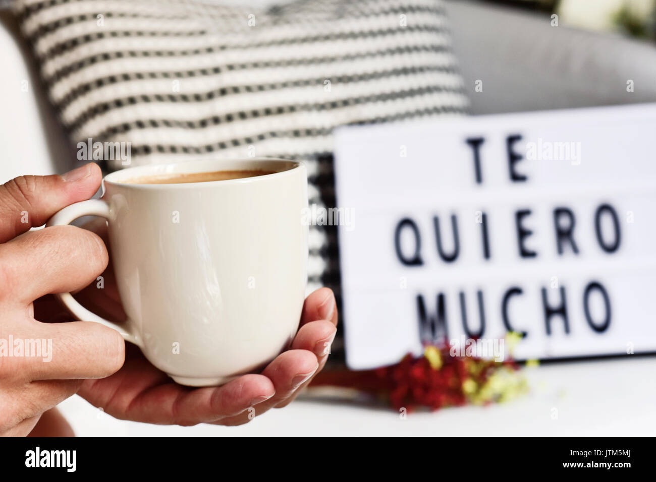 closeup of a young man with a white ceramic cup with coffee in his hand and a lightbox in the background with the text te quiero mucho, I love you so  Stock Photo