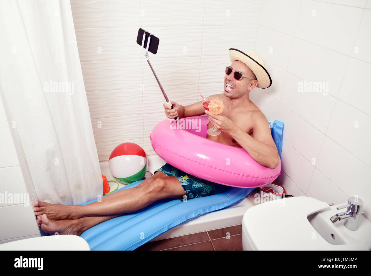 a young caucasian man wearing sunglasses, a straw hat and a pink swim ring in a blue air mattress placed in the shower of a bathroom takes a selfie wi Stock Photo