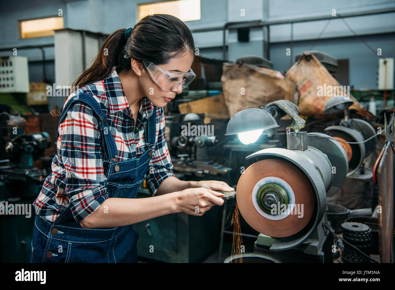 attractive lathe female worker holding components metal grinding and wearing safety glasses working on grinding wheel with sparks in milling machine w Stock Photo
