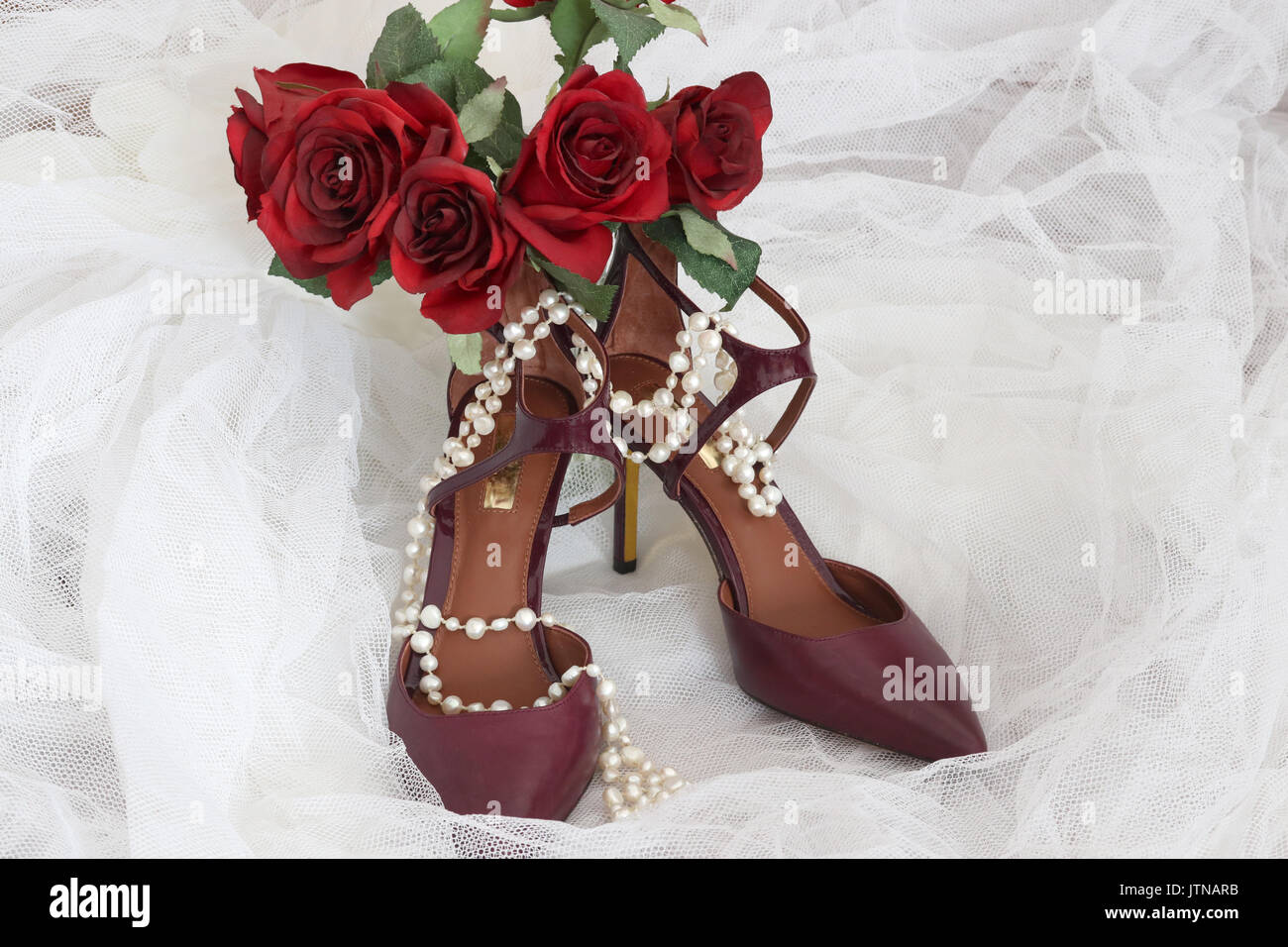 Bright shoes for stylish bride. Burgundy color high hill pair of stylish shoes and brides bouquet with red roses on a white bridal veil. Stock Photo