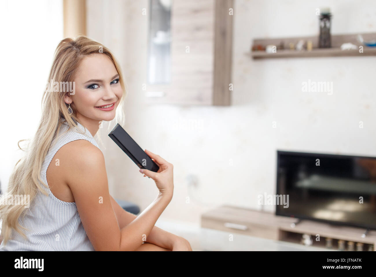 Happy blonde young woman with TV remote control in living room Stock Photo