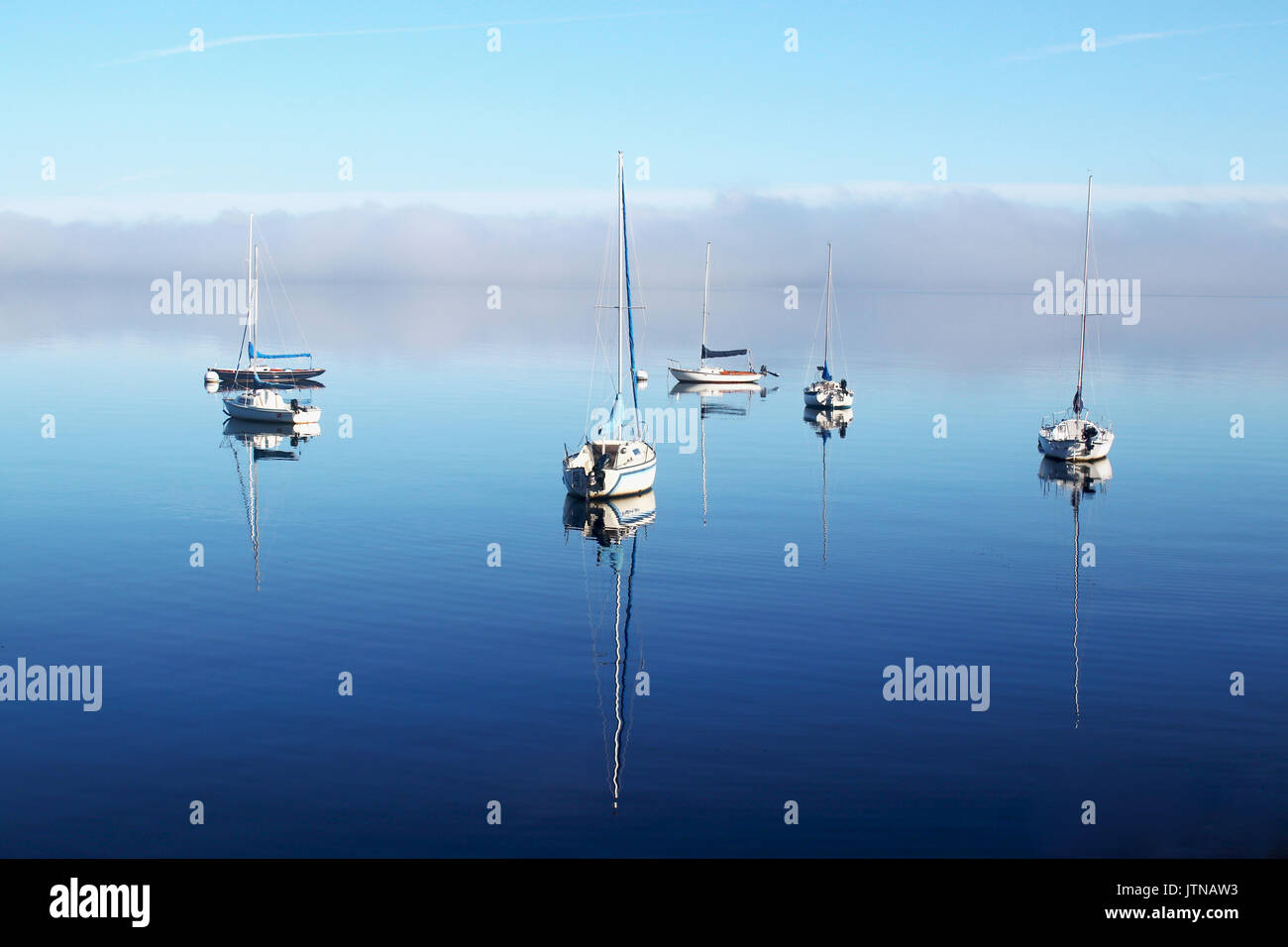 Beautiful misty morning sunrise. Landscape with a sailing boats reflected in a calm surface of the lake and a fog over waterfront. Horizontal composit Stock Photo