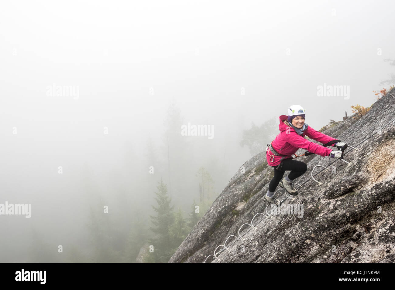 A women smiles with excitement as she climbs up the metal rungs of a Via Ferrata in Squamish. Stock Photo