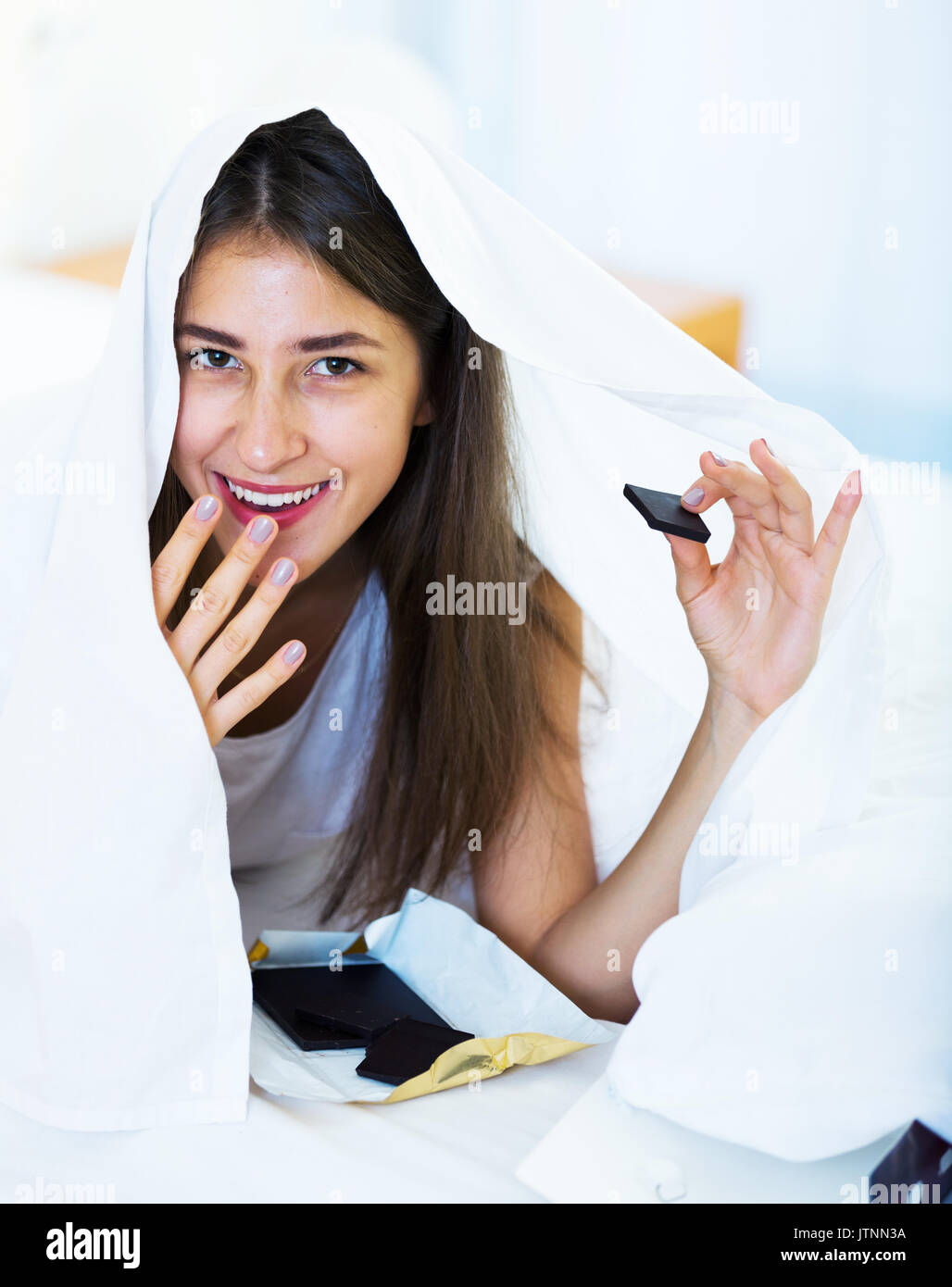 Smiling girl secretely eating chocolate under blanket in bed Stock Photo