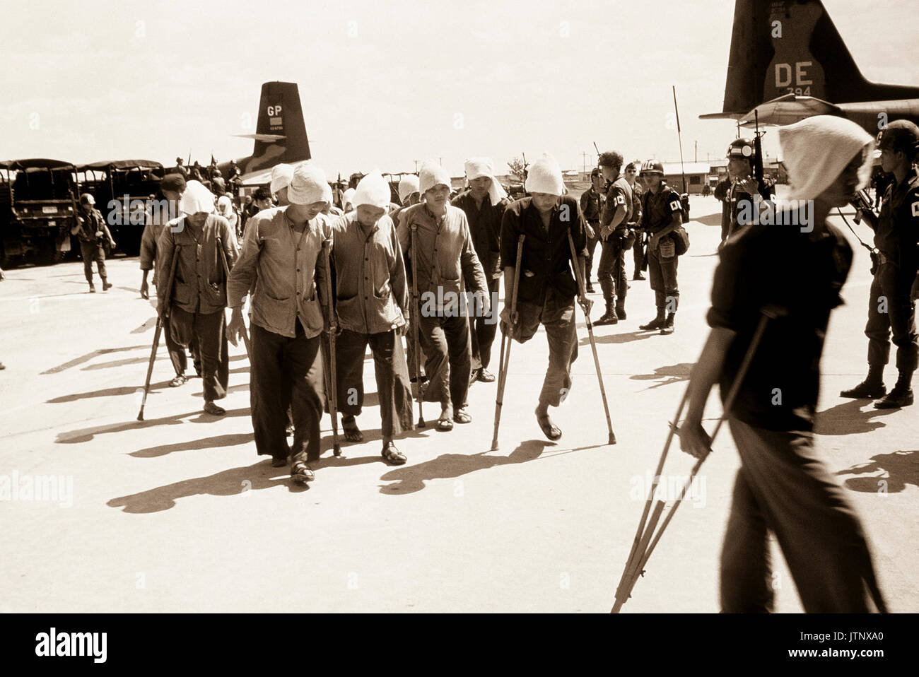 Viet Cong POWs, some on crutches, under the watchful eyes of South Vietnamese military police walk to the waiting C-123 transport aircraft.  The POWS will be airlifted to Loc Ninh, South Vietnam for the prisoner exchange between the United States/South Vietnam and North Vietnam/Viet Cong militaries. Stock Photo