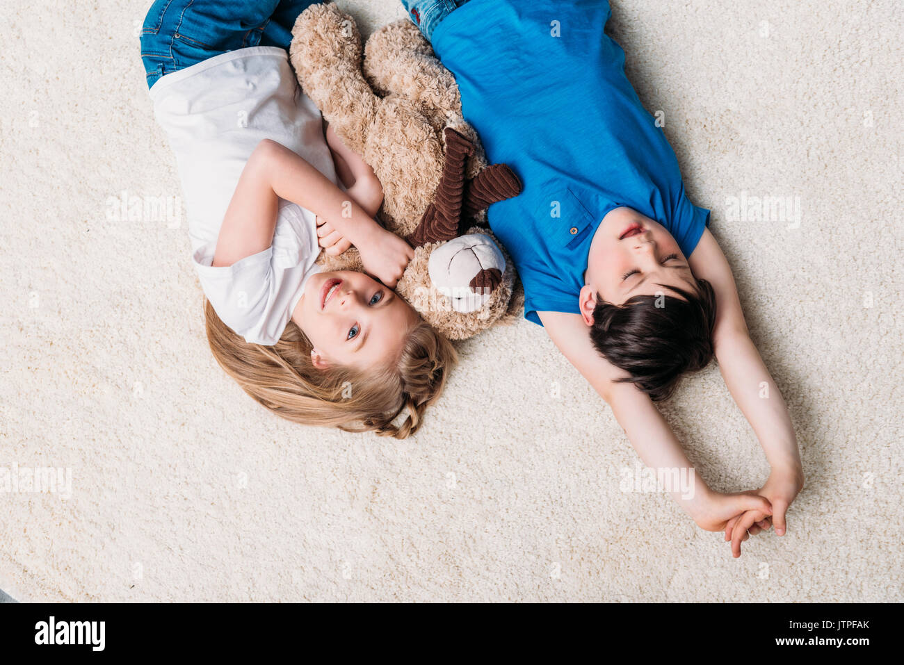 little boy and girl lying on carpet with teddy bear at home and looking at camera Stock Photo