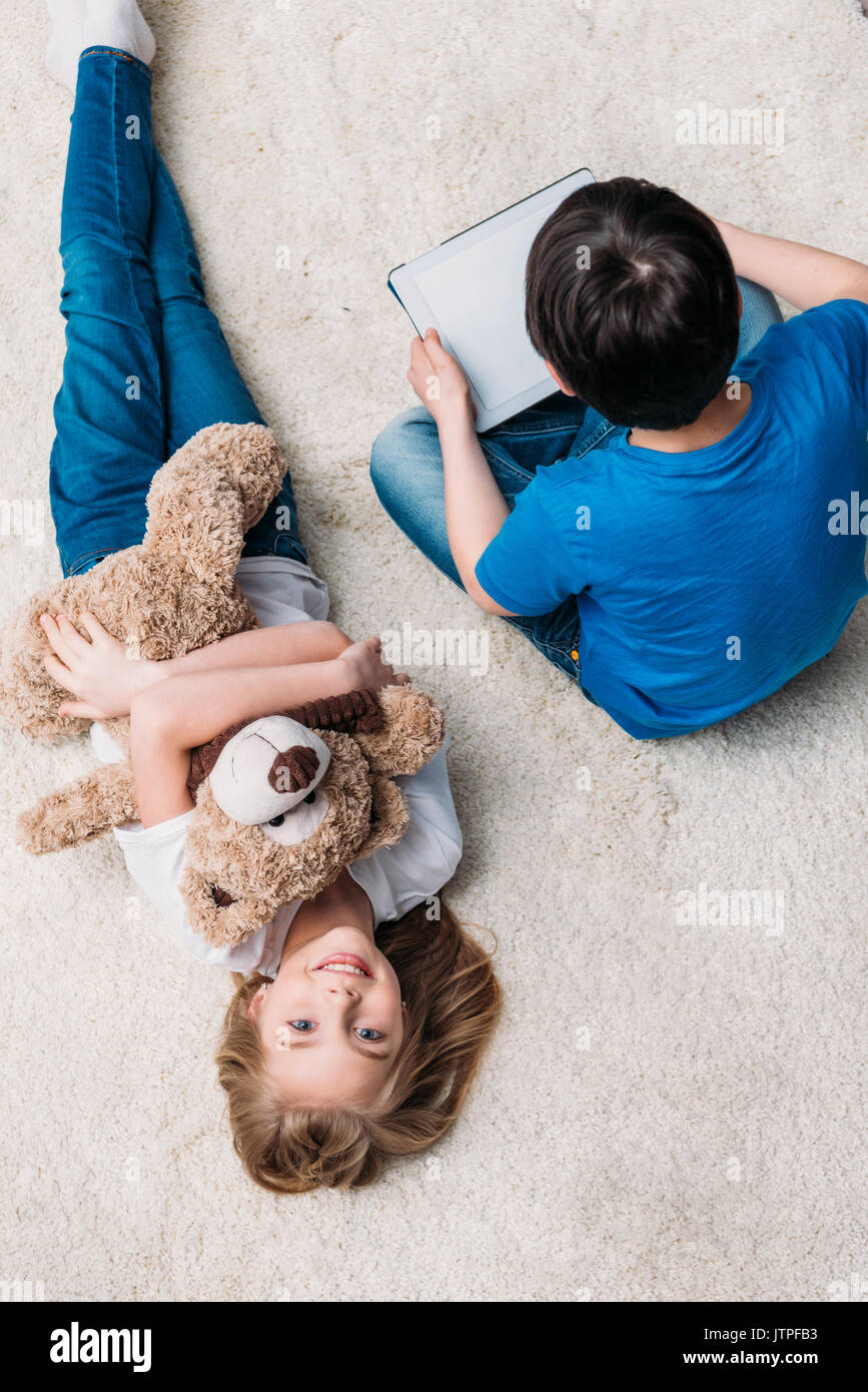 little girl with teddy bear and boy with digital tablet on carpet at home Stock Photo