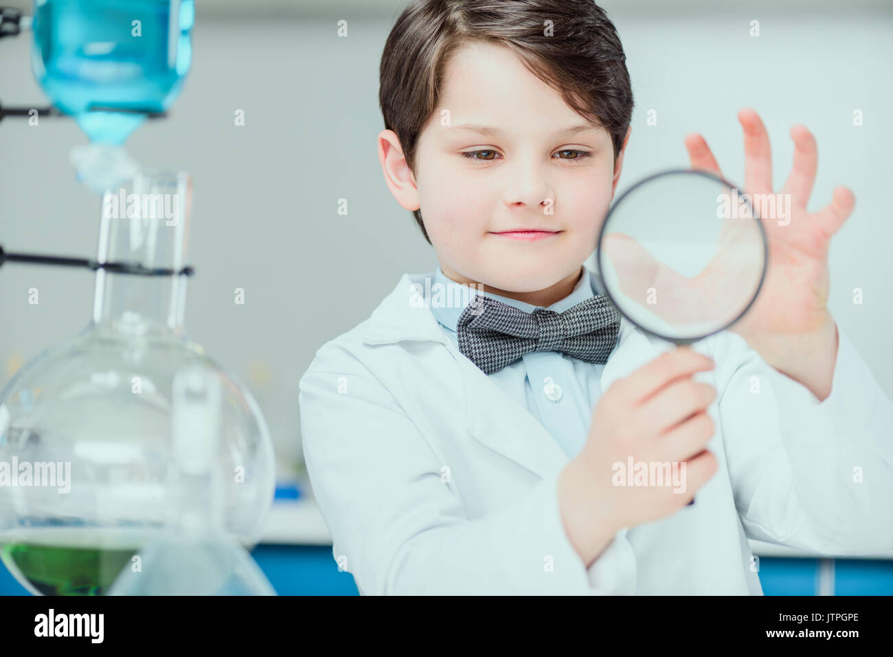 Little scientist in white coat holding magnifier in chemical lab, science school concept Stock