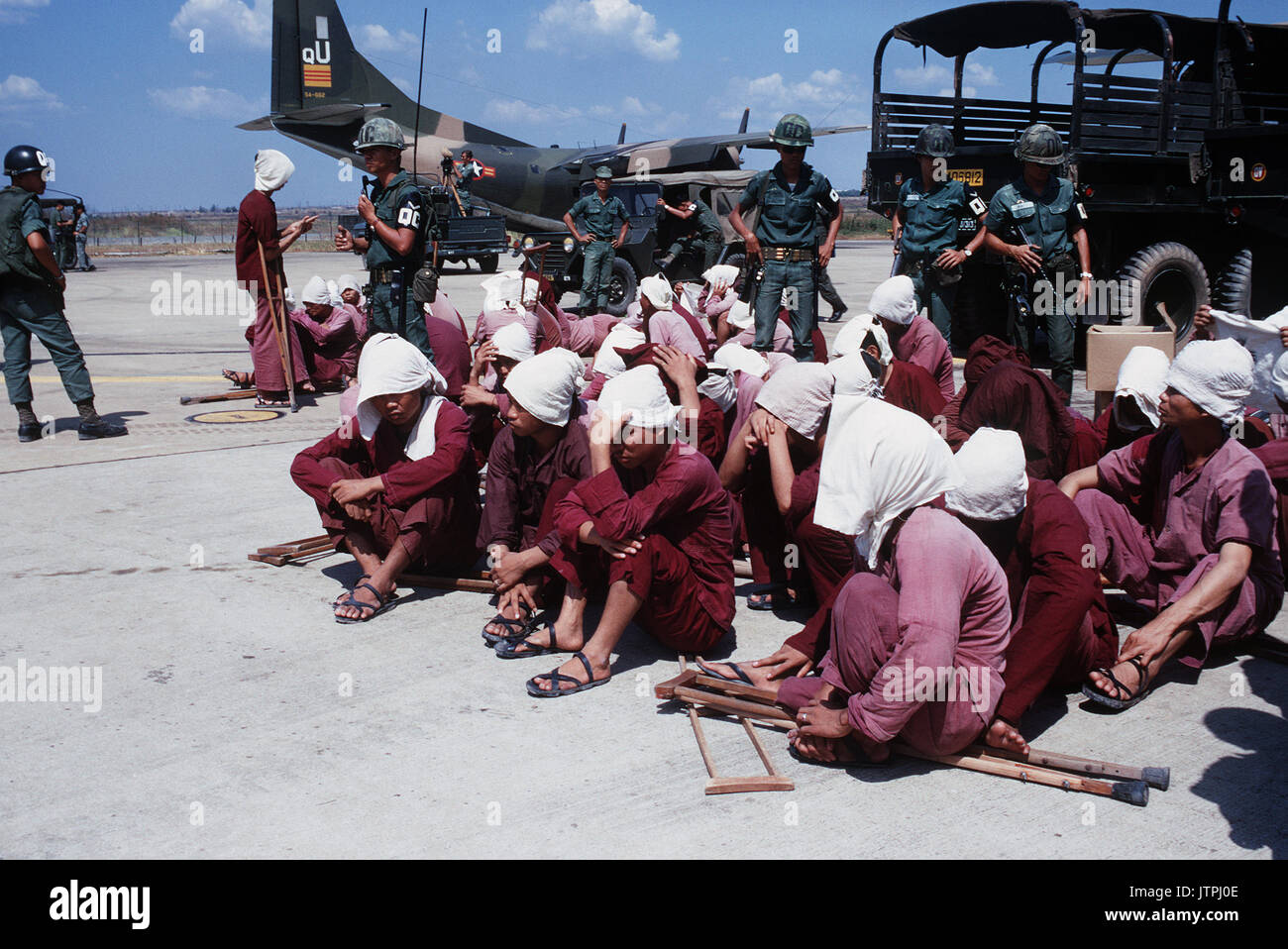 Viet Cong POWs sit on the ramp at Tan Son Nhut Air Base under the watchful eyes of South Vietnamese military police.  The POWs were brought to the airbase in the 6X6 trucks in the background and will be airlifted to Loc Ninh, South Vietnam on the C-123 transport aircraft for the prisoner exchange between the United States/South Vietnam and North Vietnam/Viet Cong militaries. Stock Photo