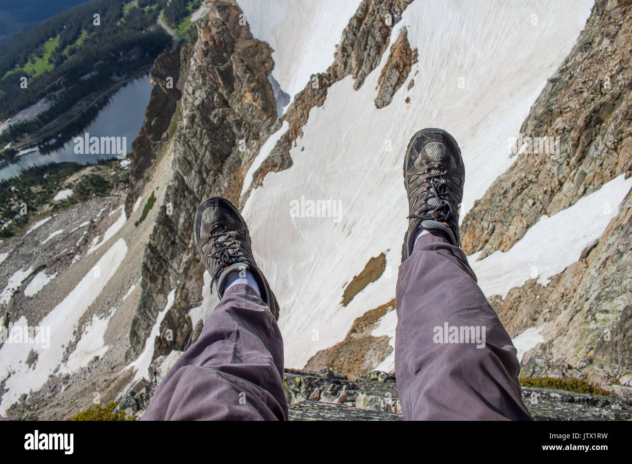 A hiker dangles his legs over the edge of a cliff in the Rocky Mountains. Stock Photo
