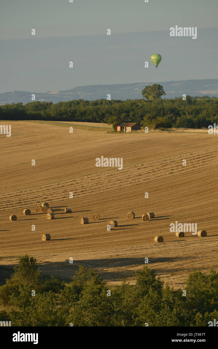 Hot air balloon (Montgolfier) over a French rural landscape in summer. Hay bales on harvested brown fields. Near Cesteyrols, Occitanie, France. Stock Photo