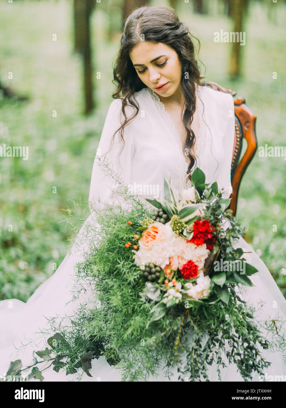 The portrait of the woman with the brown hair in the white long wedding dress sitting on the chair and holding at looking at the huge bouquet of red and white flowers in the forest. Stock Photo