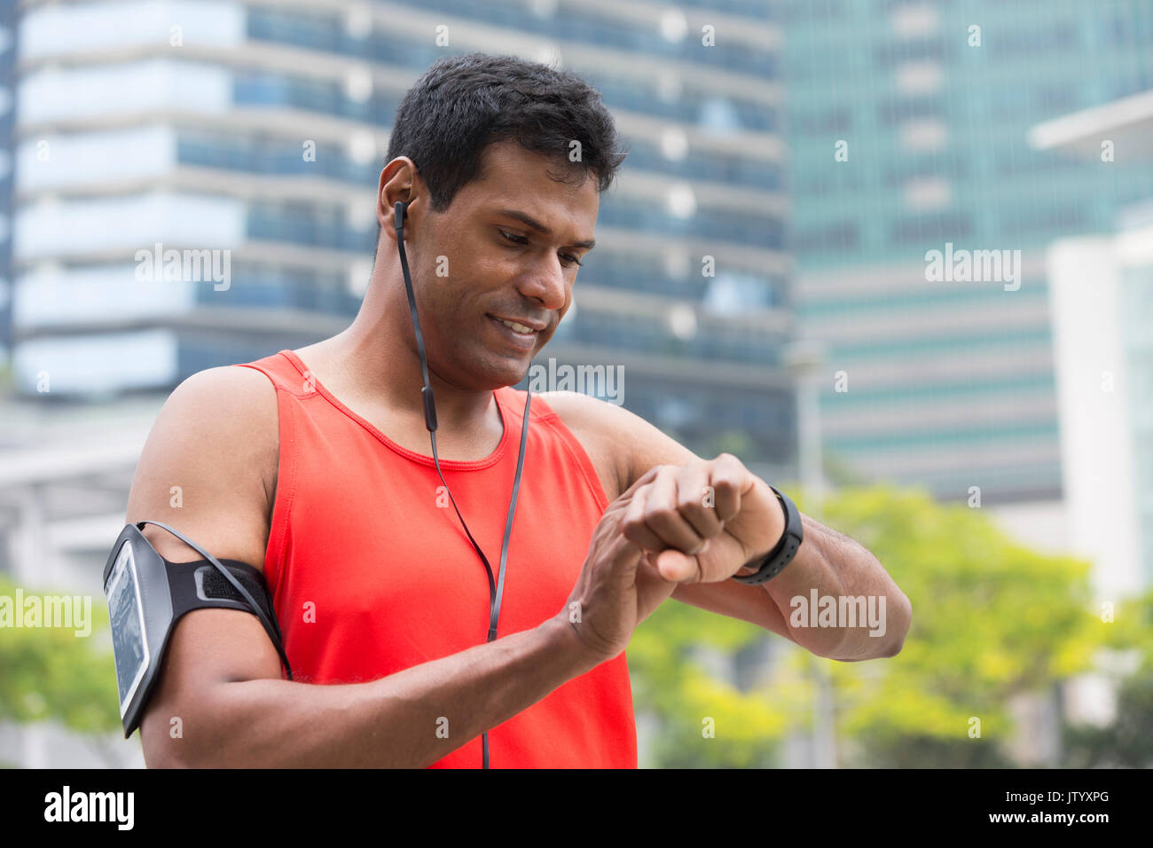Sporty Indian man looking at his smartwatch heart rate monitor. Athletic Asian man using a smart watch and listening to music with earphones. Stock Photo