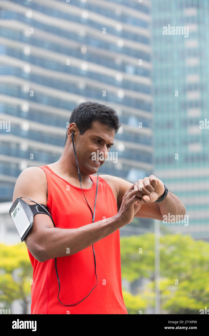 Sporty Indian man looking at his smartwatch heart rate monitor. Athletic Asian man using a smart watch and listening to music with earphones. Stock Photo