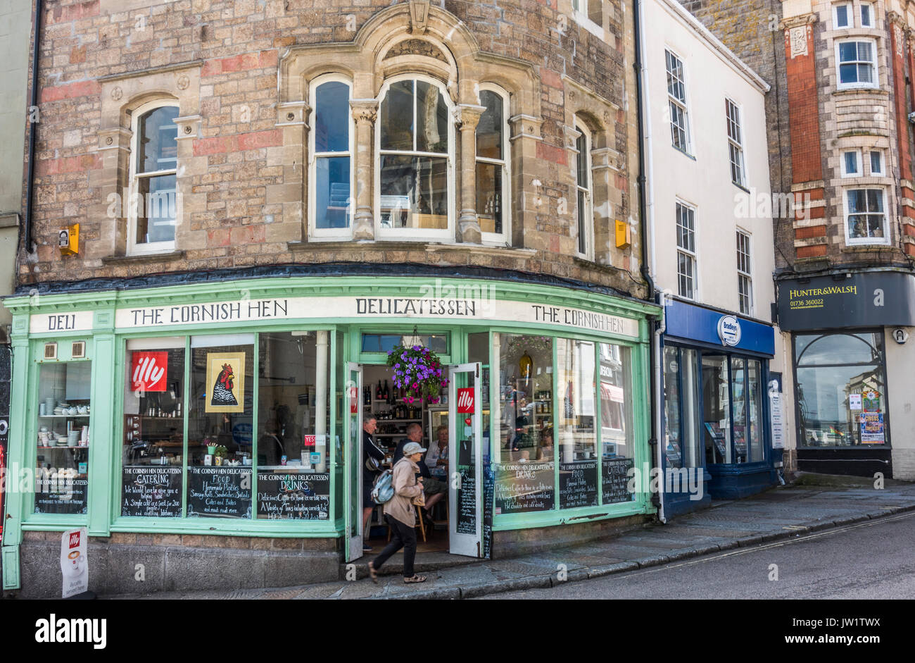 Lady walking past the open doors of The Cornish Hen deli shop, in the centre of Penzance, Cornwall, England, UK. Stock Photo