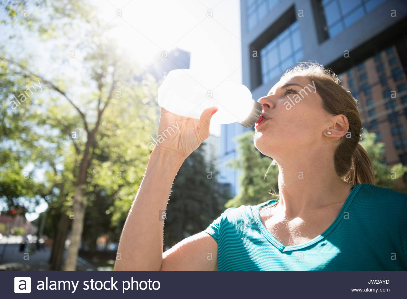 Female runner drinking water from water bottle in sunny urban park Stock Photo
