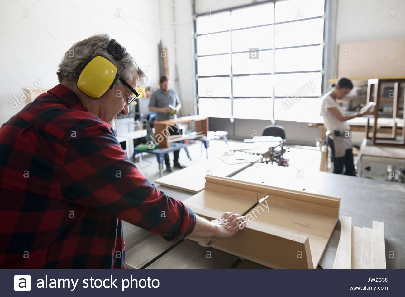 Male carpenter with ear protectors cutting wood at table saw in workshop Stock Photo