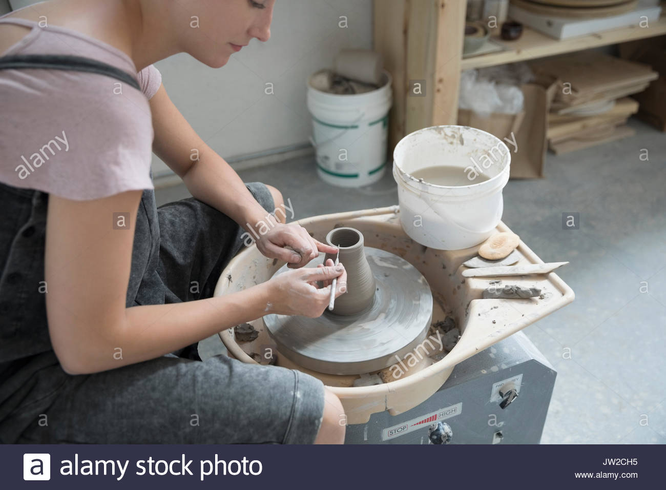 Female potter using tool to shape wet clay at pottery wheel in art