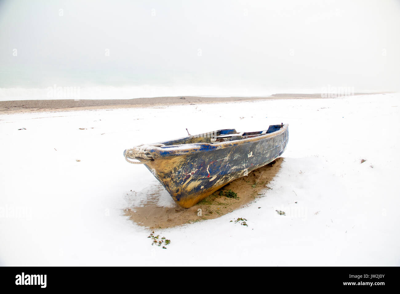 Old abandoned blue wooden boat under white snow on beach, lake covered by snow and foot traces, panoramic view of winter landscape in the beach Stock Photo