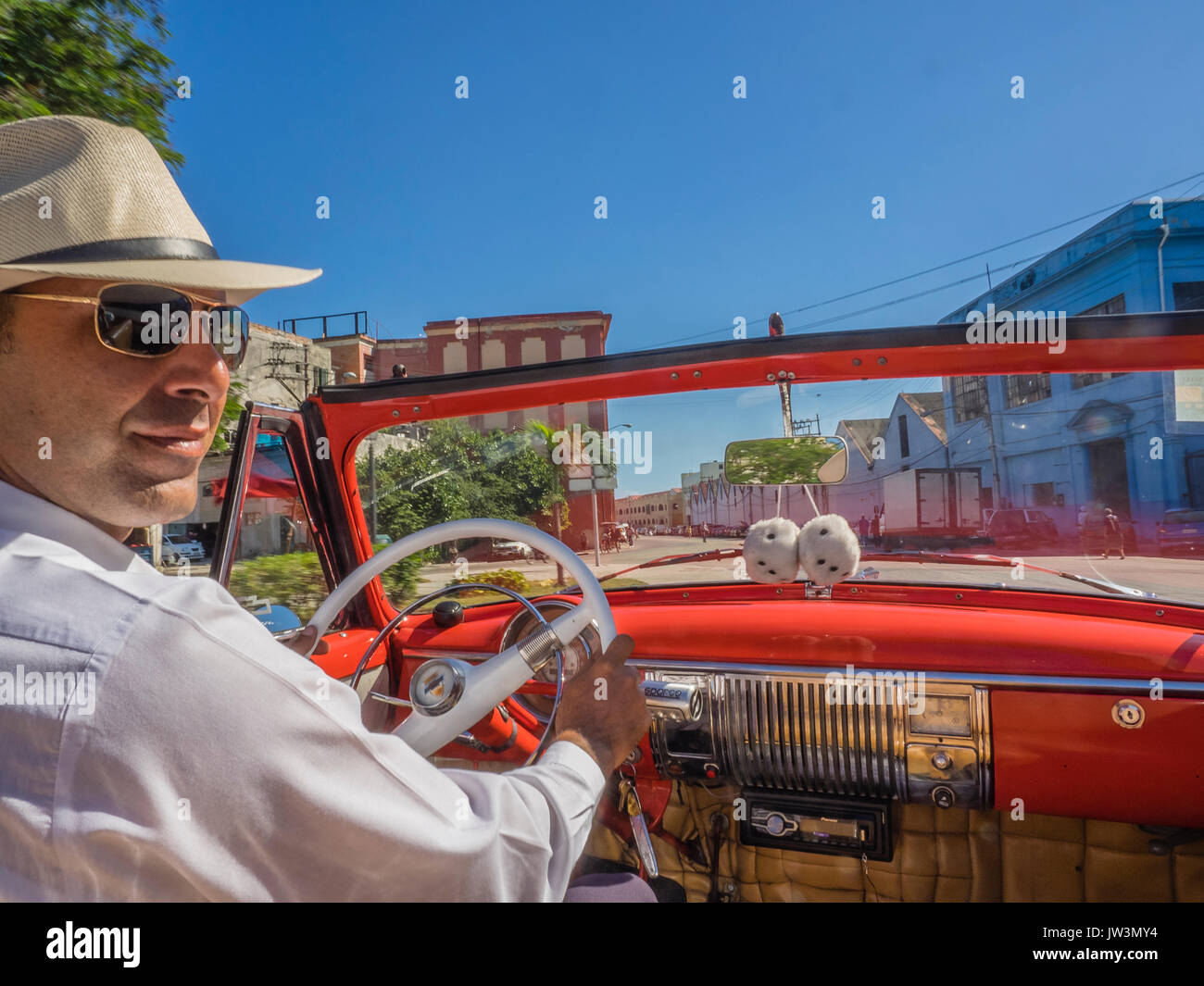 Cuban taxi driver in straw hat and white shirt at the wheel of his red 1951 Chevrolet convertible driving through the streets of old Havana with fuzzy Stock Photo