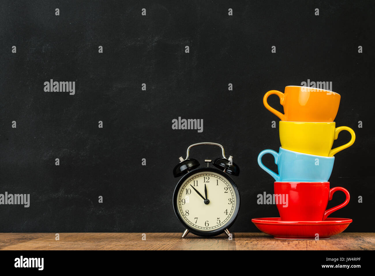 retro small alarm clock on wood texture floor over blackboard wall background in creative studio with four stacked color attractive coffee cups show d Stock Photo