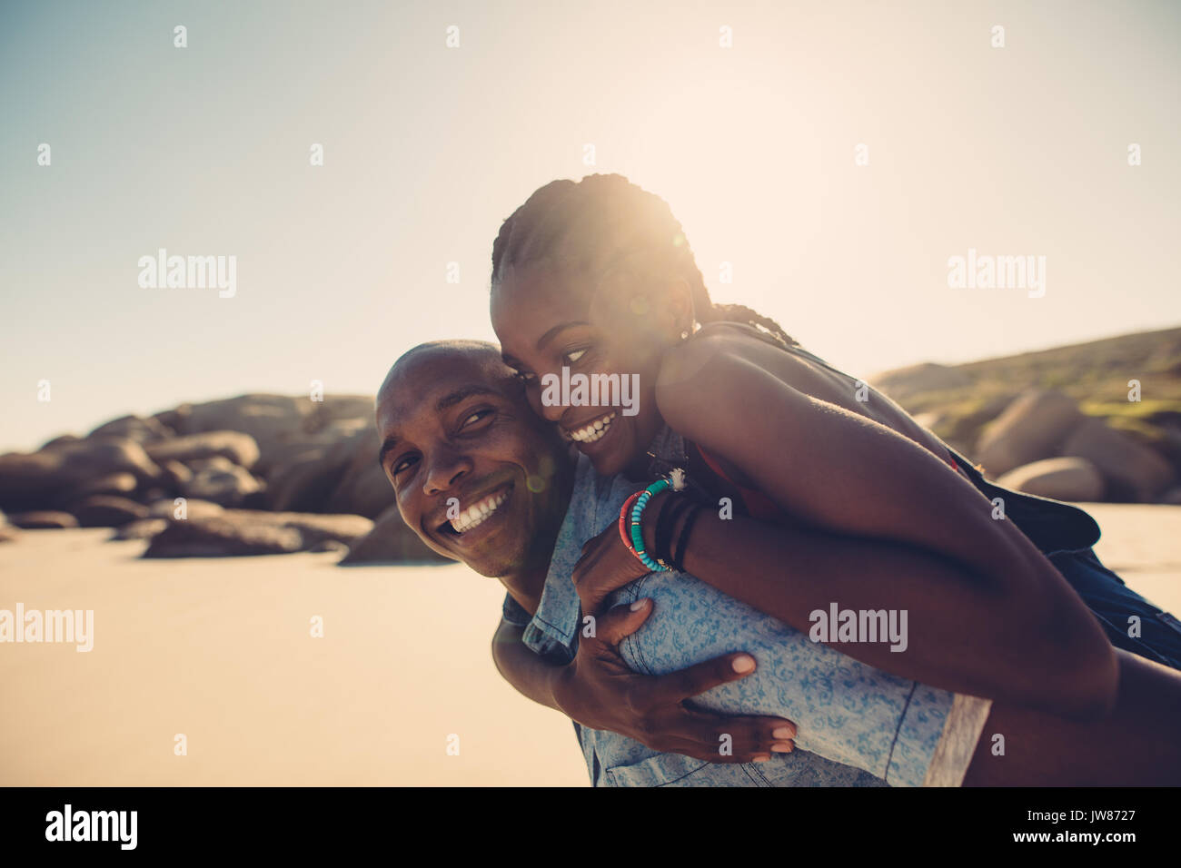 African young man carrying woman on his back at the beach. Boyfriend giving piggyback ride to his beautiful girlfriend at seashore on a summer day. Stock Photo