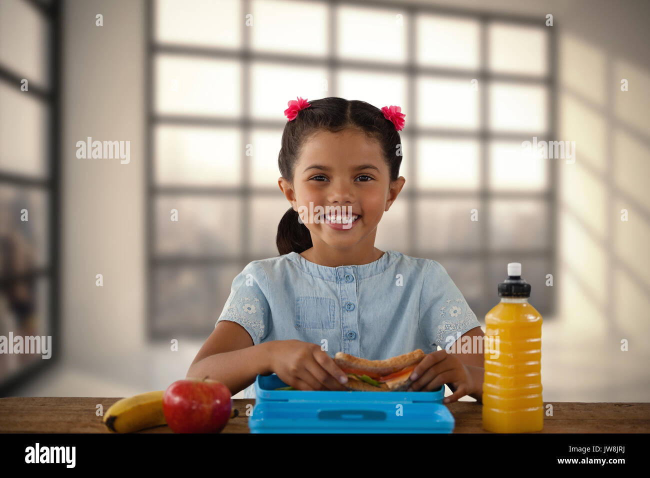 Smiling girl having sandwich at table against room with large window showing city Stock Photo
