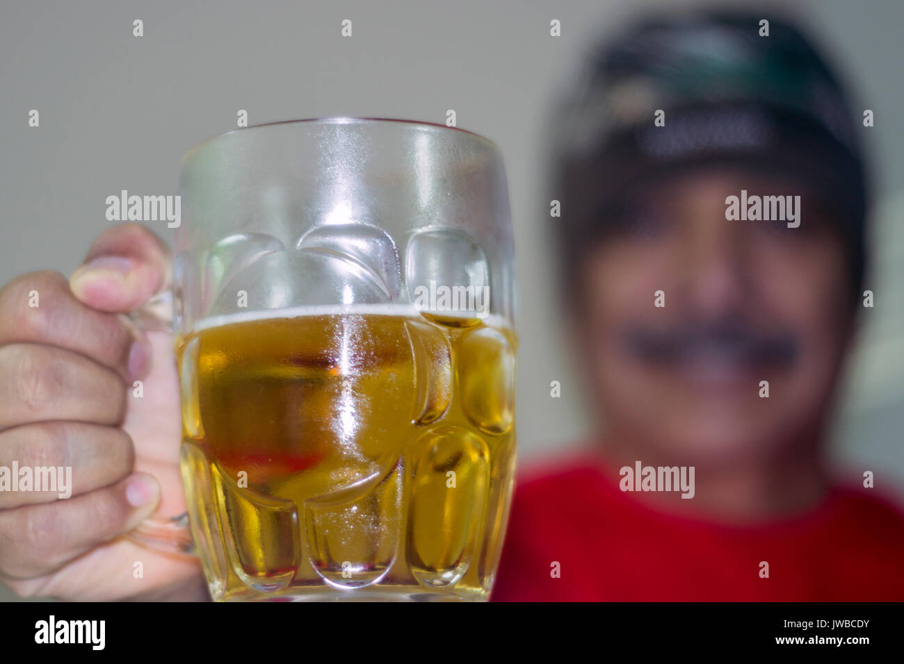 Vaso de cerveza en primer plano, un hombre desenfocado atras, empuñando el vaso, saludando y sonriendo, toma horizontal Stock Photo