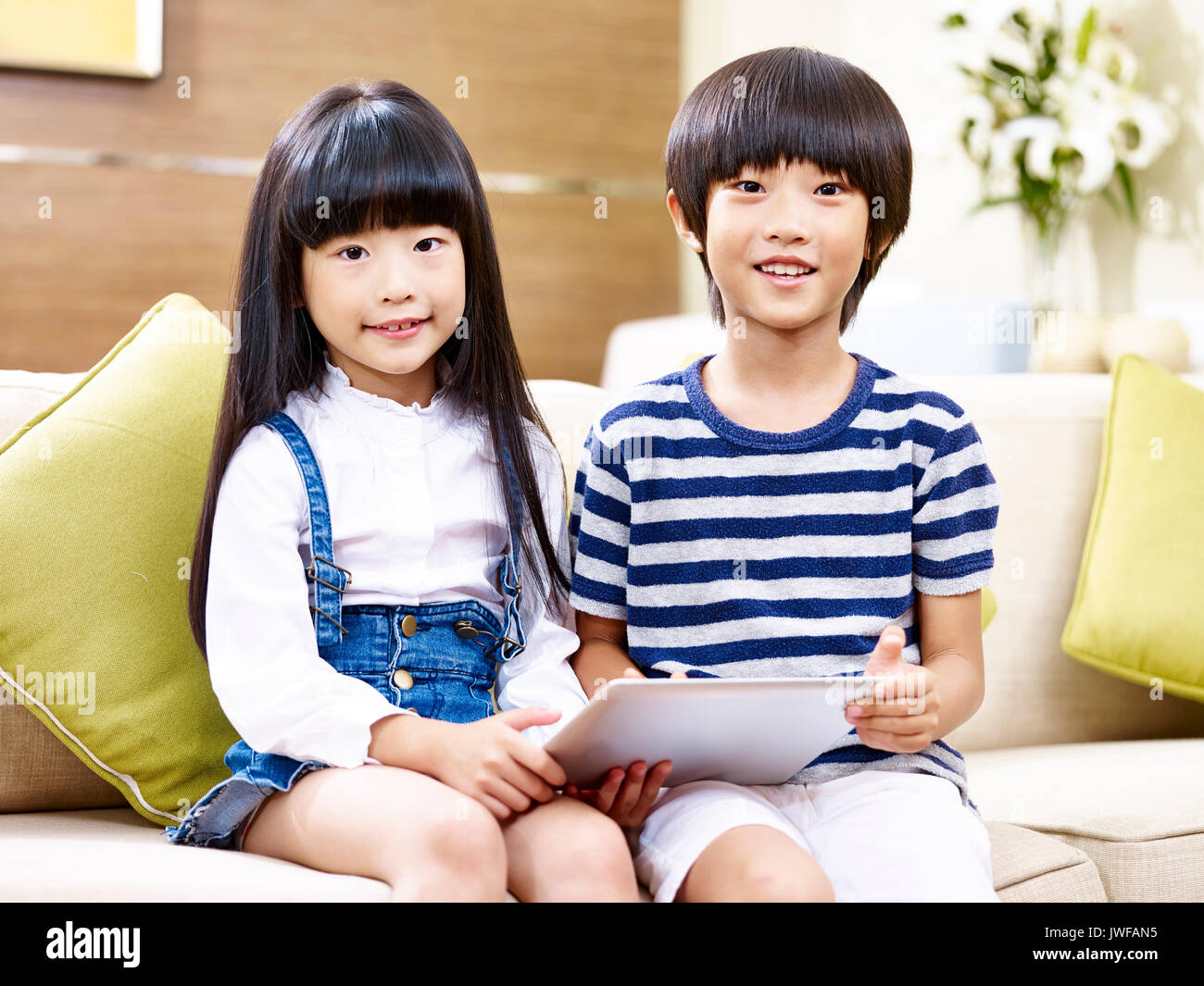 portrait of two asian children sitting on couch holding digital tablet at home. Stock Photo