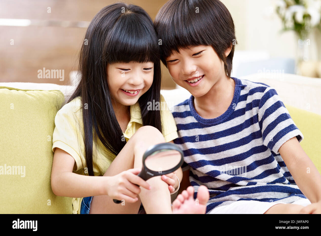 two asian children sitting on couch at home having fun playing with a magnifying glass. Stock Photo